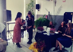 Fotografia barwna. Na pierwszym planie kobieta wręczająca dyplom uznania dziewczynie w sukience, obok znajdują się siedzące  przy stoliku trzy dziewczyny.