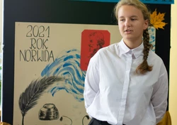 Fotografia barwna, na pierwszym planie młoda kobieta, w tle dekoracja z napisem „konkurs recytatorski”, ”2021 rok Norwida” oraz rysunek pióra z kałamarzem.