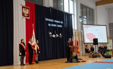 Zdjęcie przedstawia po lewej stronie poczet sztandarowy ze sztandarem szkoły, w tle flaga polski i godło, z przodu mównica na której przemawia dyrektor szkoły