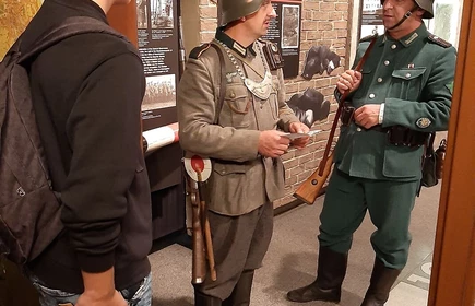 Fotografia barwna. Wnętrze muzealne. Na pierwszym planie o lewej stronie młodzieniec, po prawej dwóch mężczyzn w strojach niemieckich żołnierzy z czasów II wojny światowej. W tle fragmenty ekspozycji muzealnej.