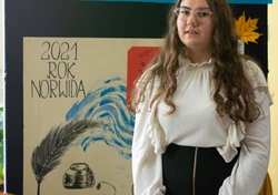 Fotografia barwna, na pierwszym planie młoda kobieta z długimi, ciemnymi włosami. W tle widoczna dekoracja z napisem „konkurs recytatorski”, „2021 rok Norwida” oraz rysunek pióra z kałamarzem.