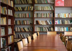 wnętrze czytelni, na pierwszym planie długi stół z krzesłami, w tle regały z książkami