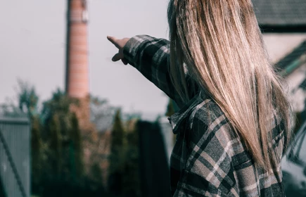 Na zdjęciu w oddali komin fabryki na pierwszym planie dziewczyna z długimi włosami odwrócona plecami i wskazująca palcem na komin