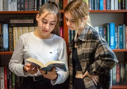 dwie dziewczyny czytajace ksiazke na tle regału z książkami. Dziewczyna po lewej w bałej bluzce trzyma ksiązkę, dziewczyna po prawej w koszuli w kratkę