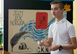 Fotografia barwna, na pierwszym planie młody mężczyzna, w tle dekoracja z napisem „konkurs recytatorski”.