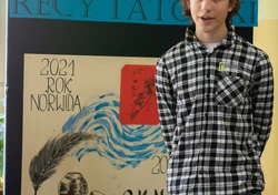 Fotografia barwna, na pierwszym planie młody mężczyzna, w tle dekoracja z napisem „konkurs recytatorski”, ”2021 rok Norwida” oraz rysunek pióra z kałamarzem.