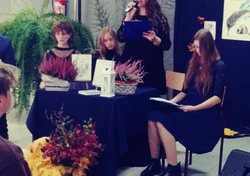 Fotografia barwna. Na pierwszym planie trzy  młode kobiety siedzące przy stoliku oraz jedna trzymająca w prawej ręce mikrofon, w lewej zaś podkładkę. Na stoliku znajdują się dwa kwiaty- wrzosy oraz mała latarnia.