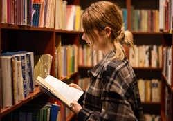 Dziewczyna stojąca lewym profilem na tle regału z książkami, w dłoniach ma otwartą książkę, dziewczyna jest w koszuli w kratkę ma bląd włosy spięte w kucyk