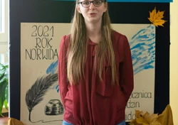Fotografia barwna, na pierwszym planie młoda kobieta z długimi, blond włosami, w tle dekoracja z napisem „konkurs recytatorski”, ”2021 rok Norwida” oraz rysunek pióra z kałamarzem.